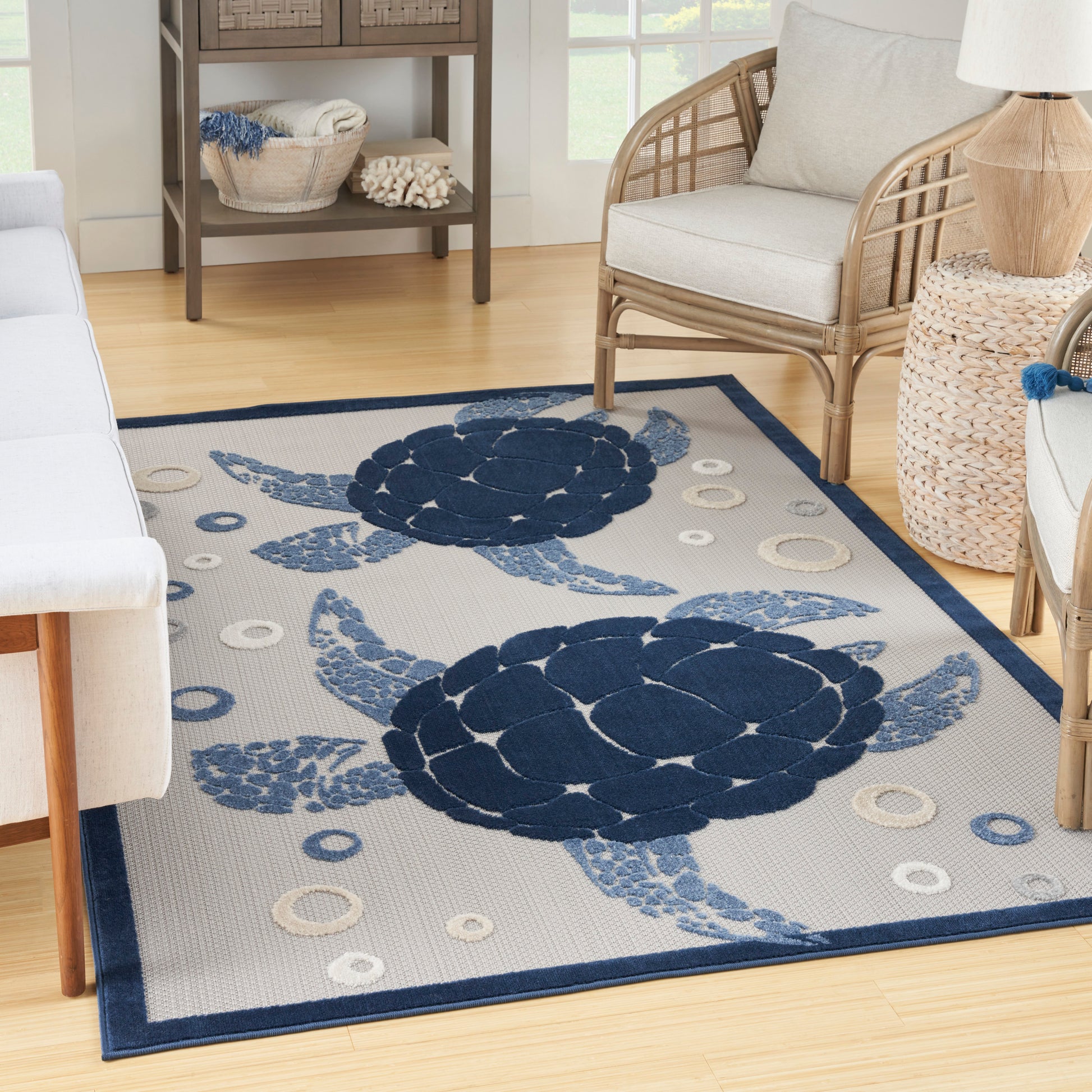 Coastal woven area rug with large navy sea turtles on a light gray field and dark blue border in a wicker seating area.