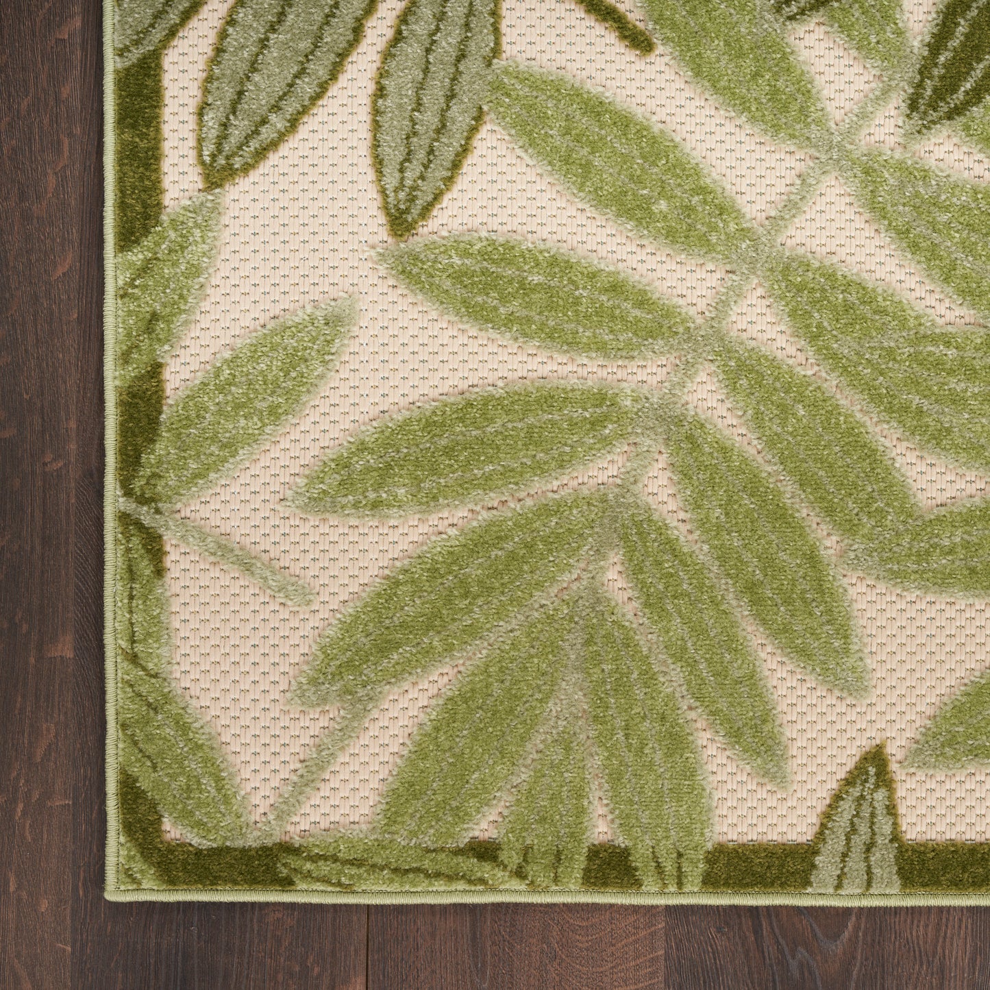 Beige area rug with large green leaf pattern, olive border, textured loop pile, on dark wood floor.