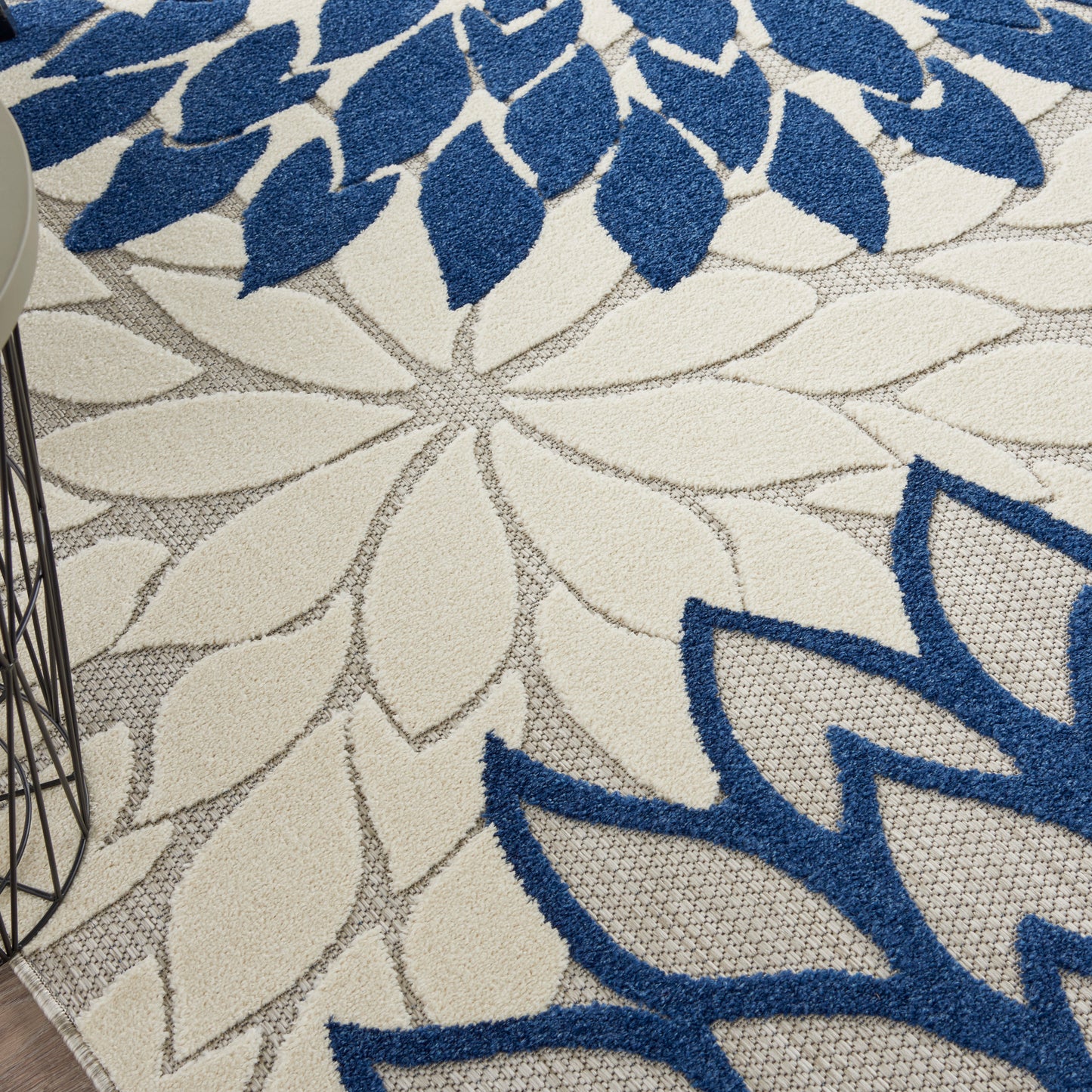 Floral area rug with large blue petals and gray outlines on cream background; partial wireframe metal side table visible.