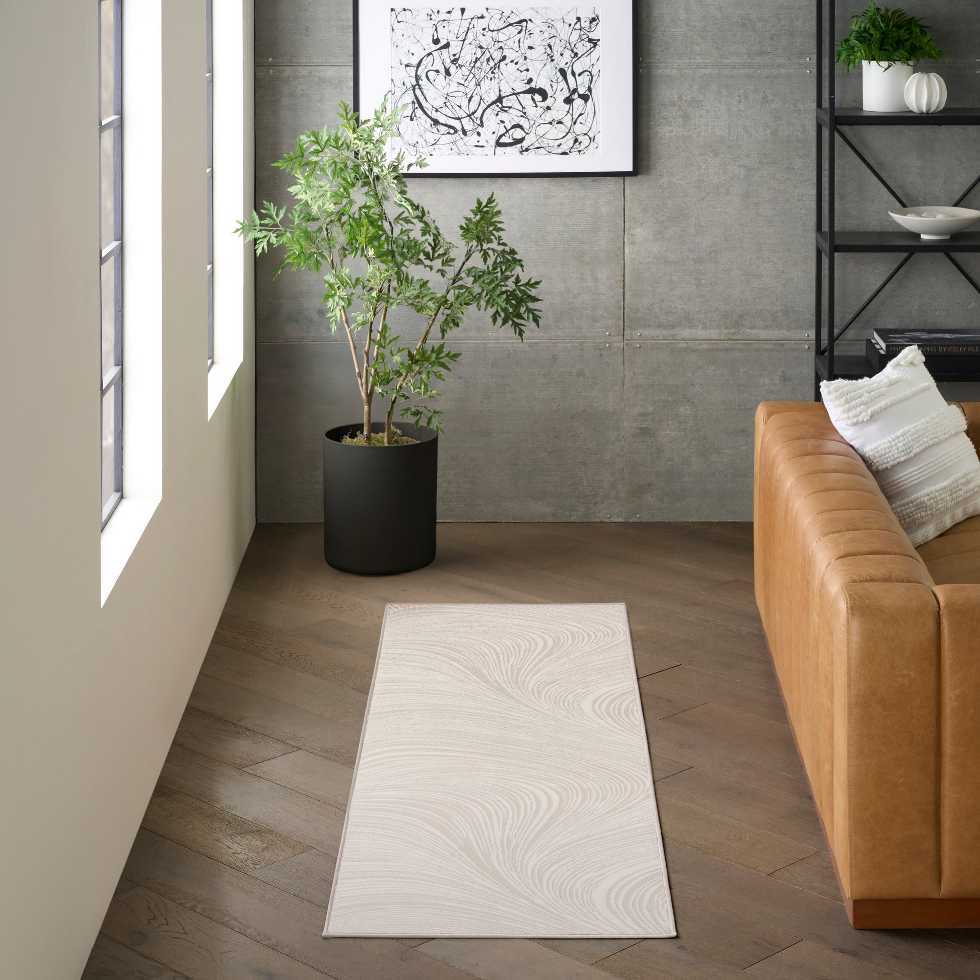 Modern living room with pale swirling runner rug, tan leather sofa, potted plant, concrete wall, abstract art & shelving.