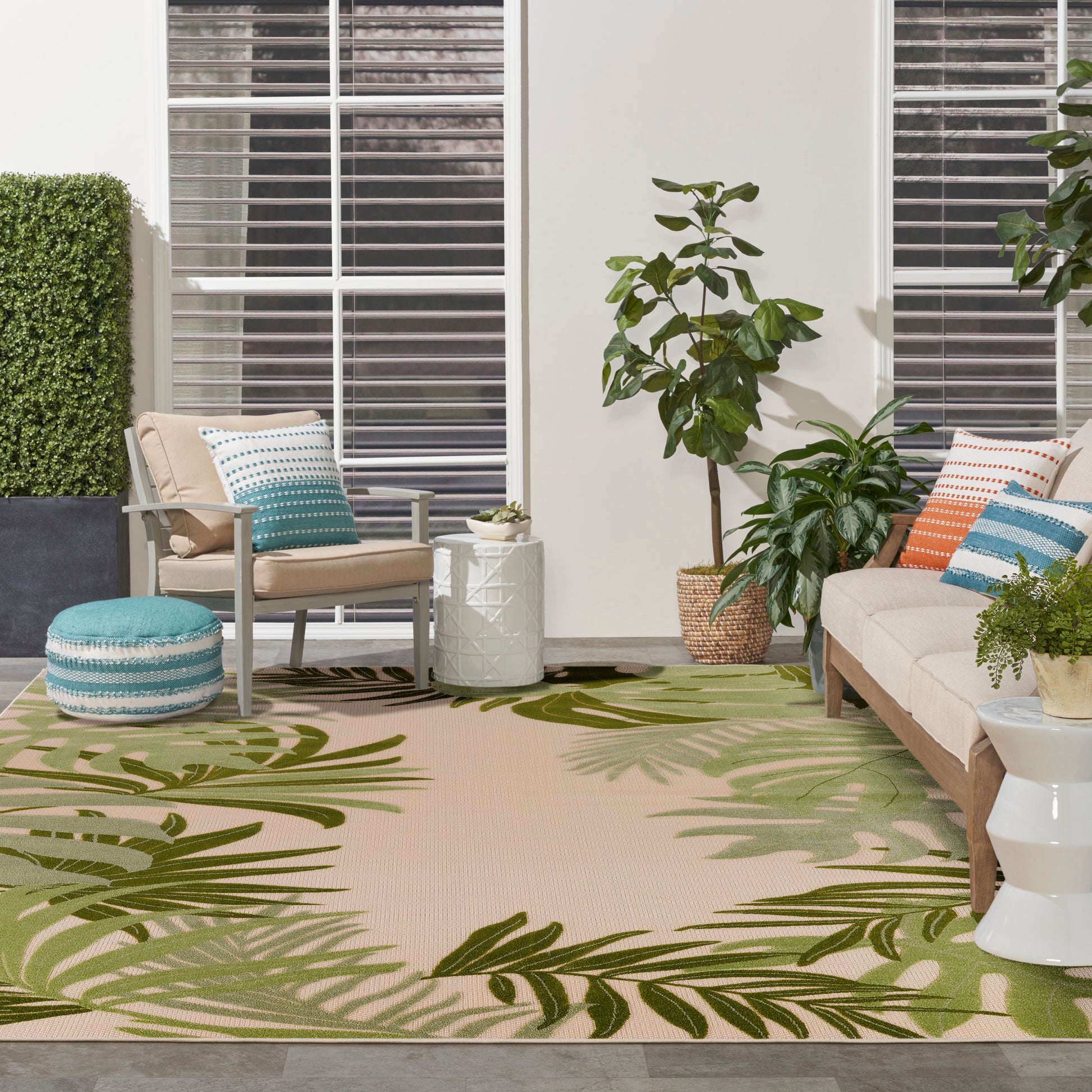 Patio lounge with palm-pattern rug, beige armchair, teal ottoman, white geometric side table, and tall potted plants.
