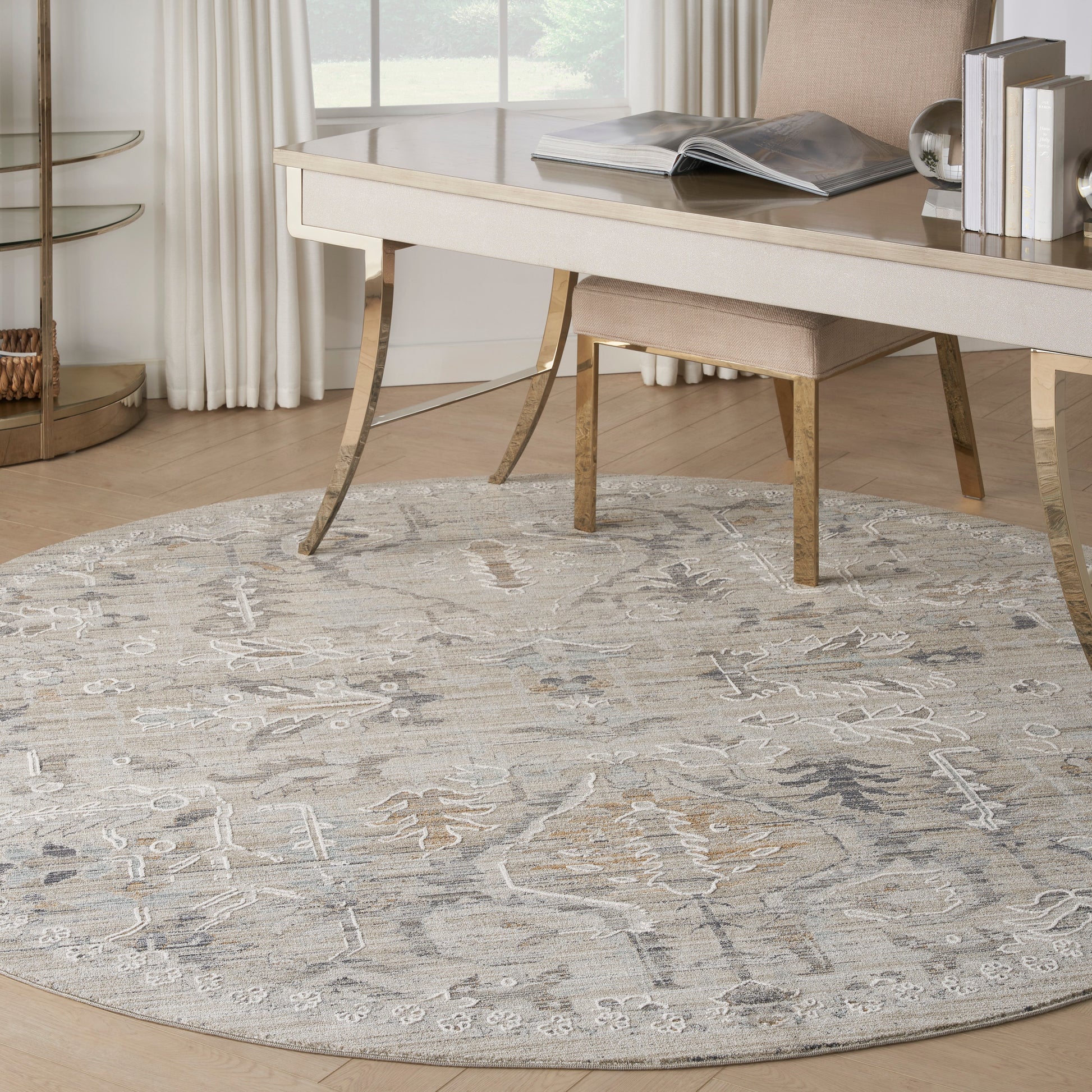 Round neutral-toned patterned area rug in gray-beige under a cream desk and light wood chair in a bright home office.