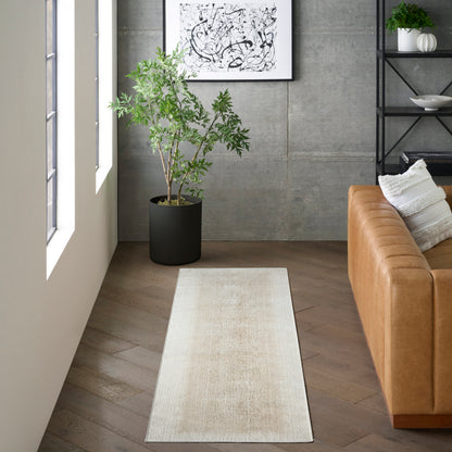 Beige runner rug with subtle texture in a modern living room; tall plant in black pot, tan leather sofa, concrete wall.