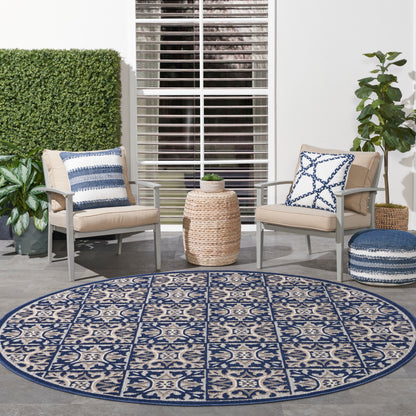 Round blue-beige rug with geometric pattern; beige cushioned chairs, a woven ottoman, and potted plants.