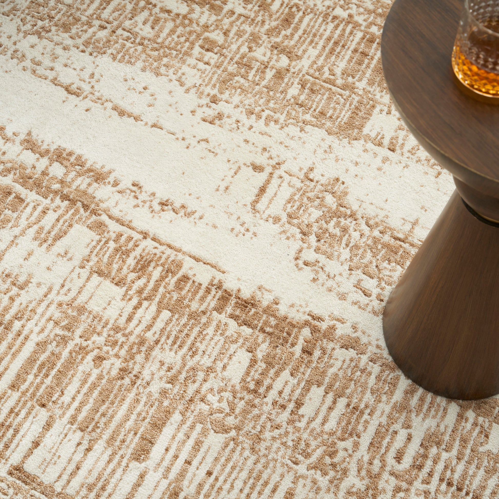 Beige-toned abstract area rug with tan stripes beside a dark wood round side table holding a glass of amber liquid.