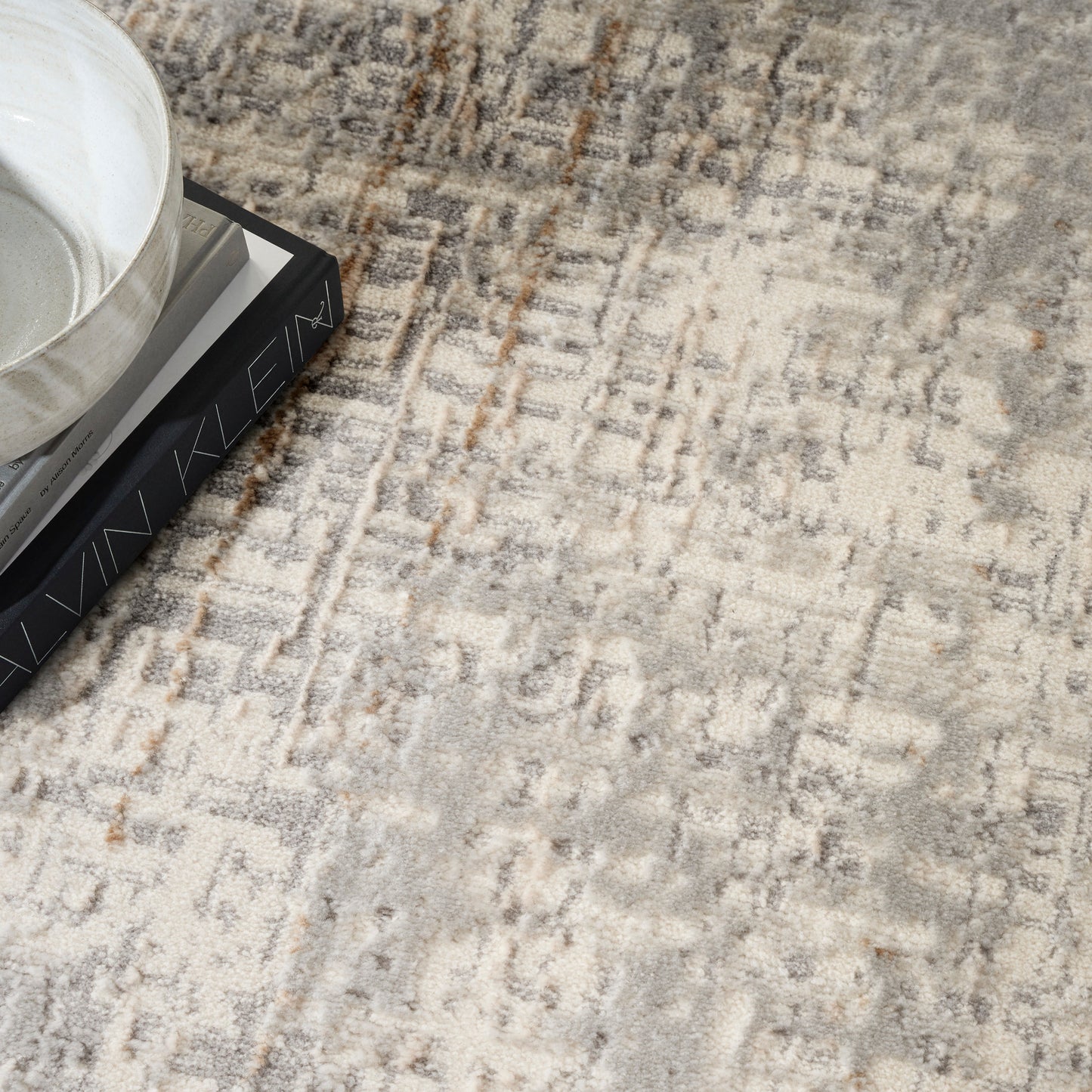 Beige-gray abstract patterned rug with a white ceramic bowl atop stacked books in the corner.
