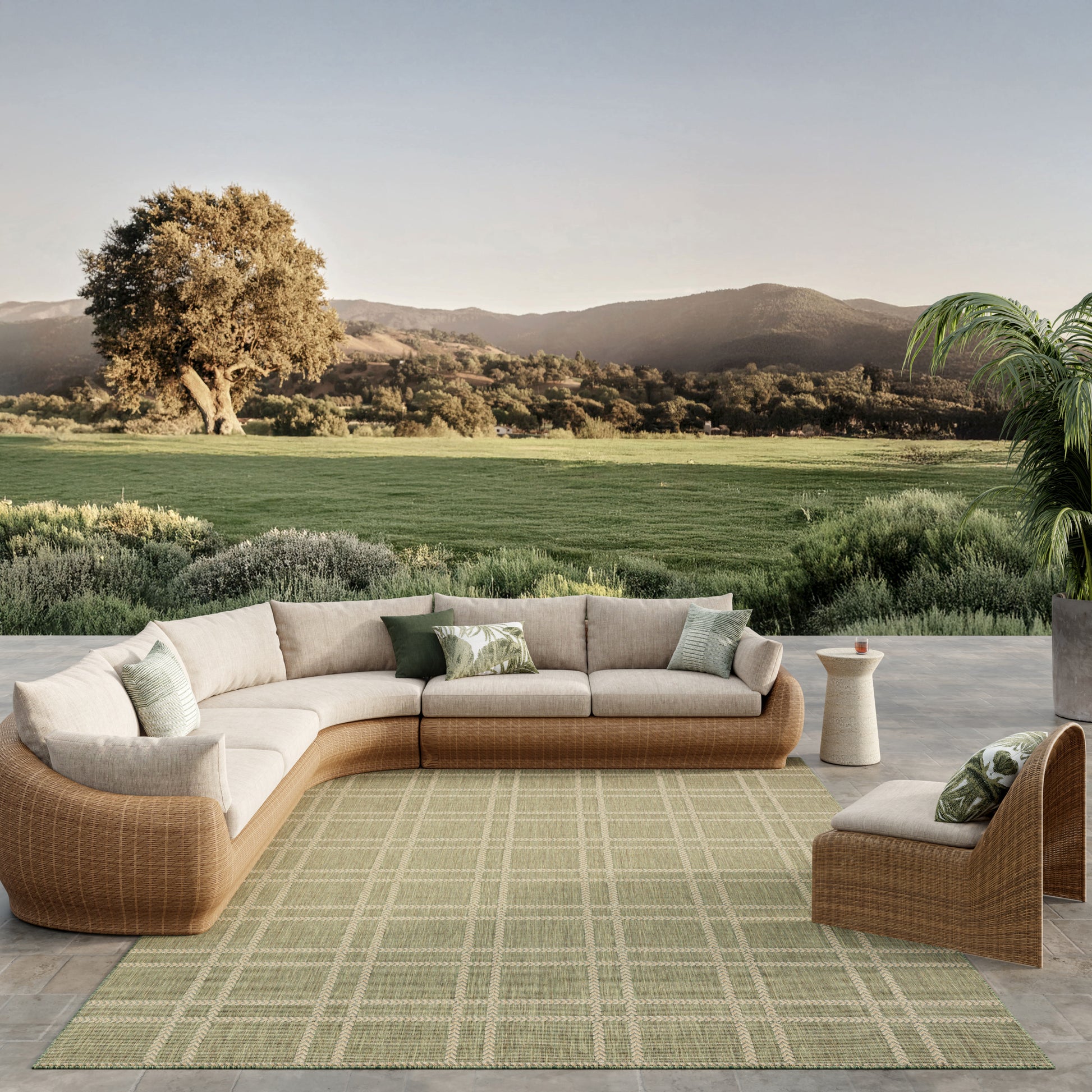 Beige woven outdoor sectional with light cushions and green pillows on a green grid rug; matching ottoman and side table.