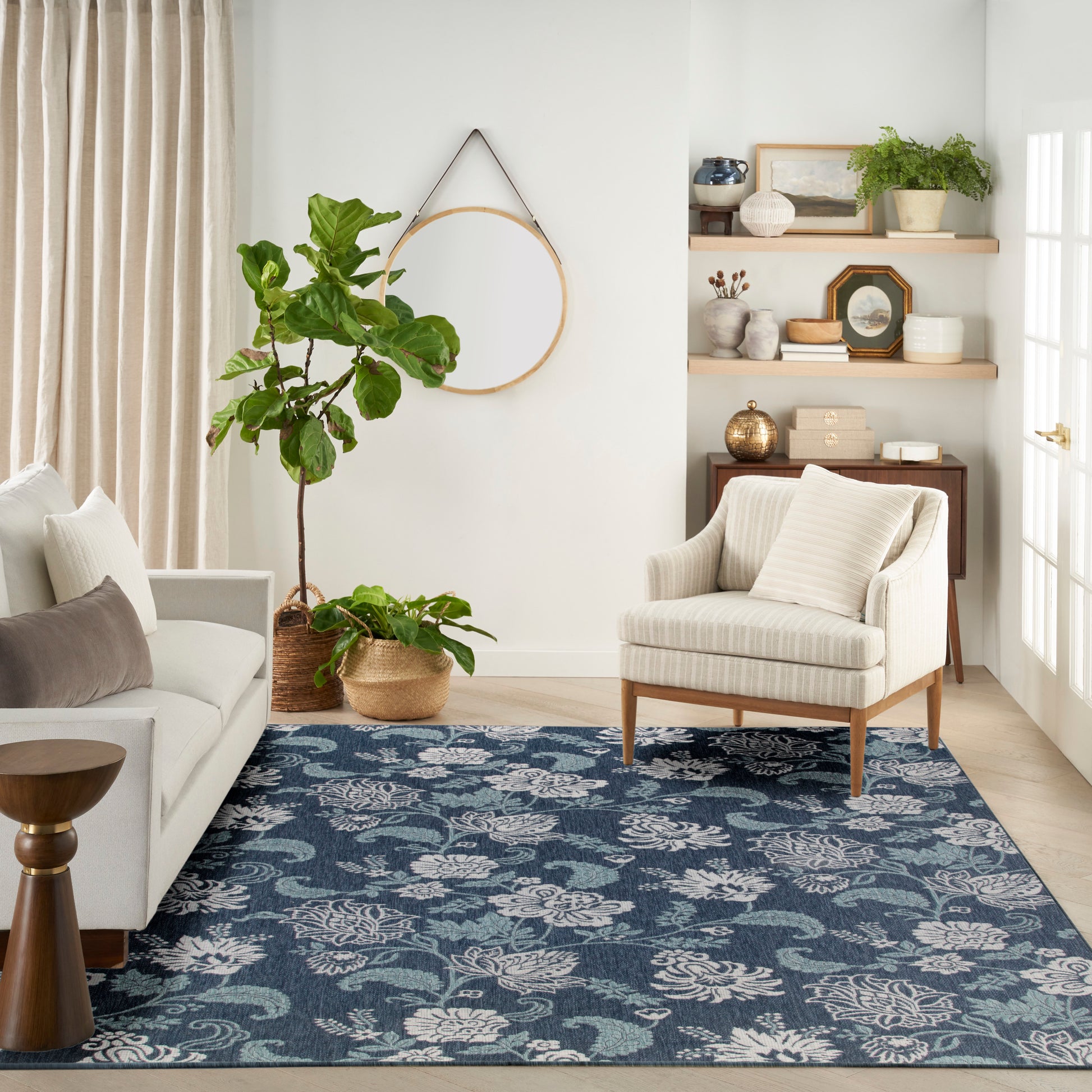 Cozy living room with blue floral rug, cream striped armchair, light sofa, wooden shelves, and large potted plants.
