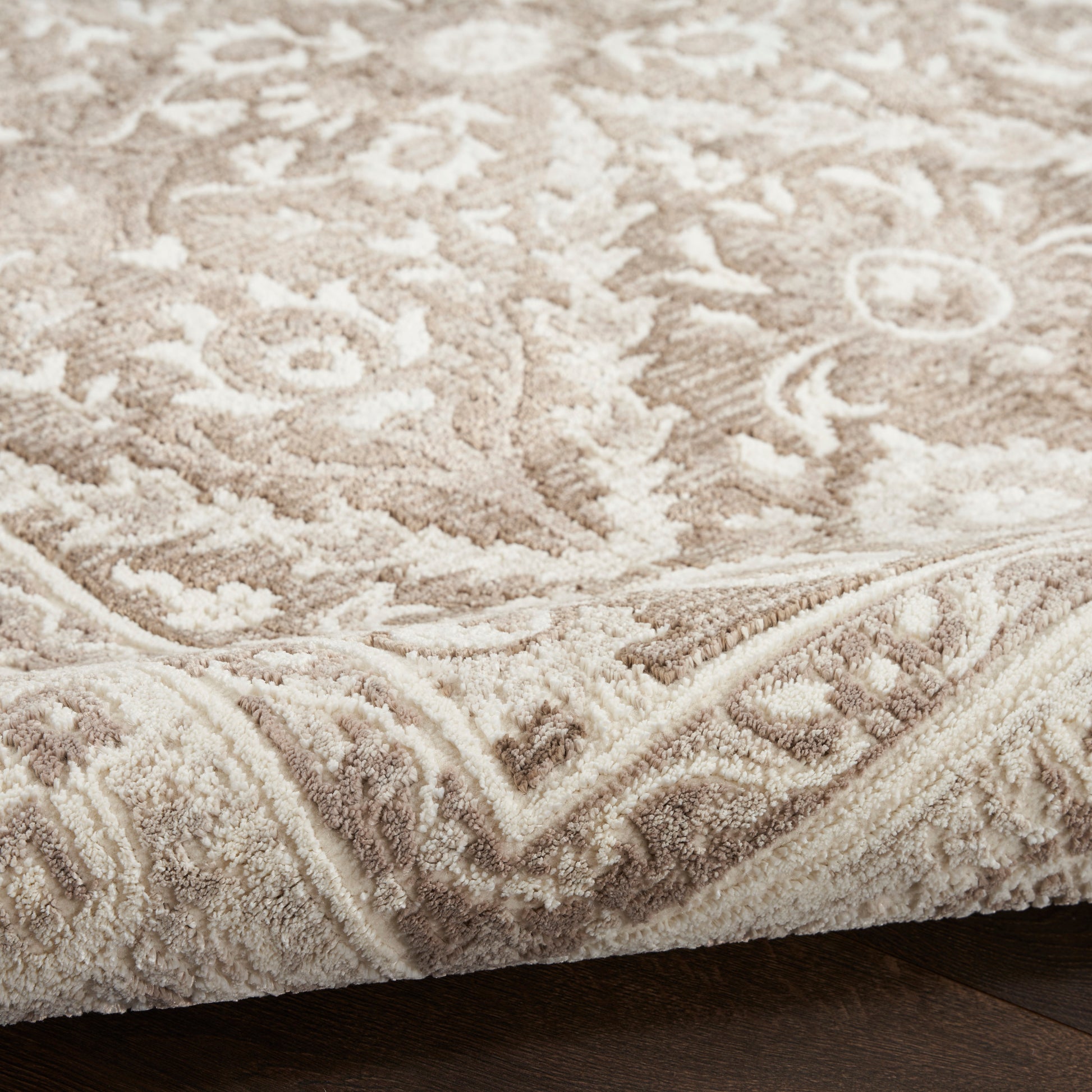 Beige ornate patterned rug with plush pile and ivory floral motifs, folded edge on dark wooden floor