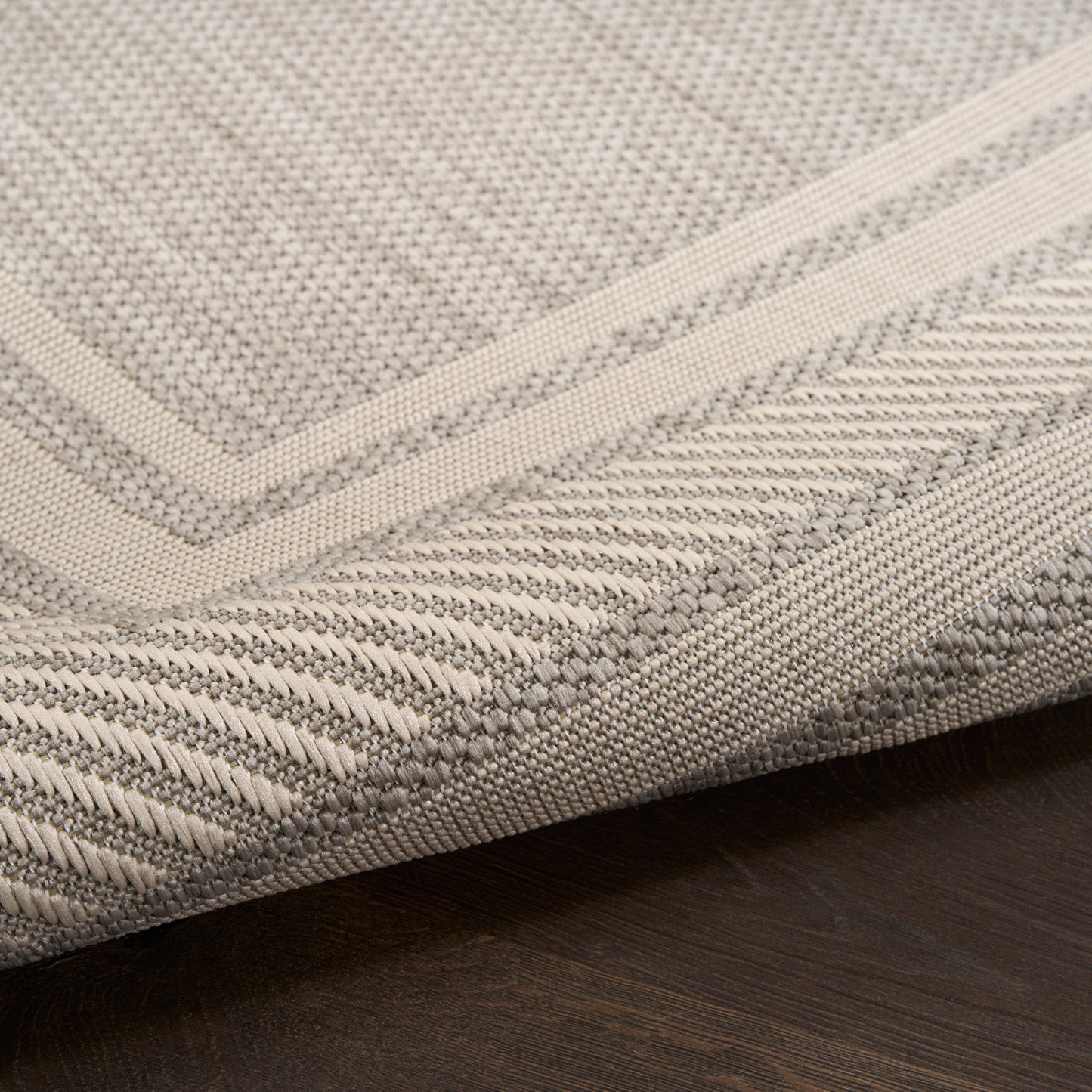Beige textured woven rug with cream border and folded corner on dark wood floor.