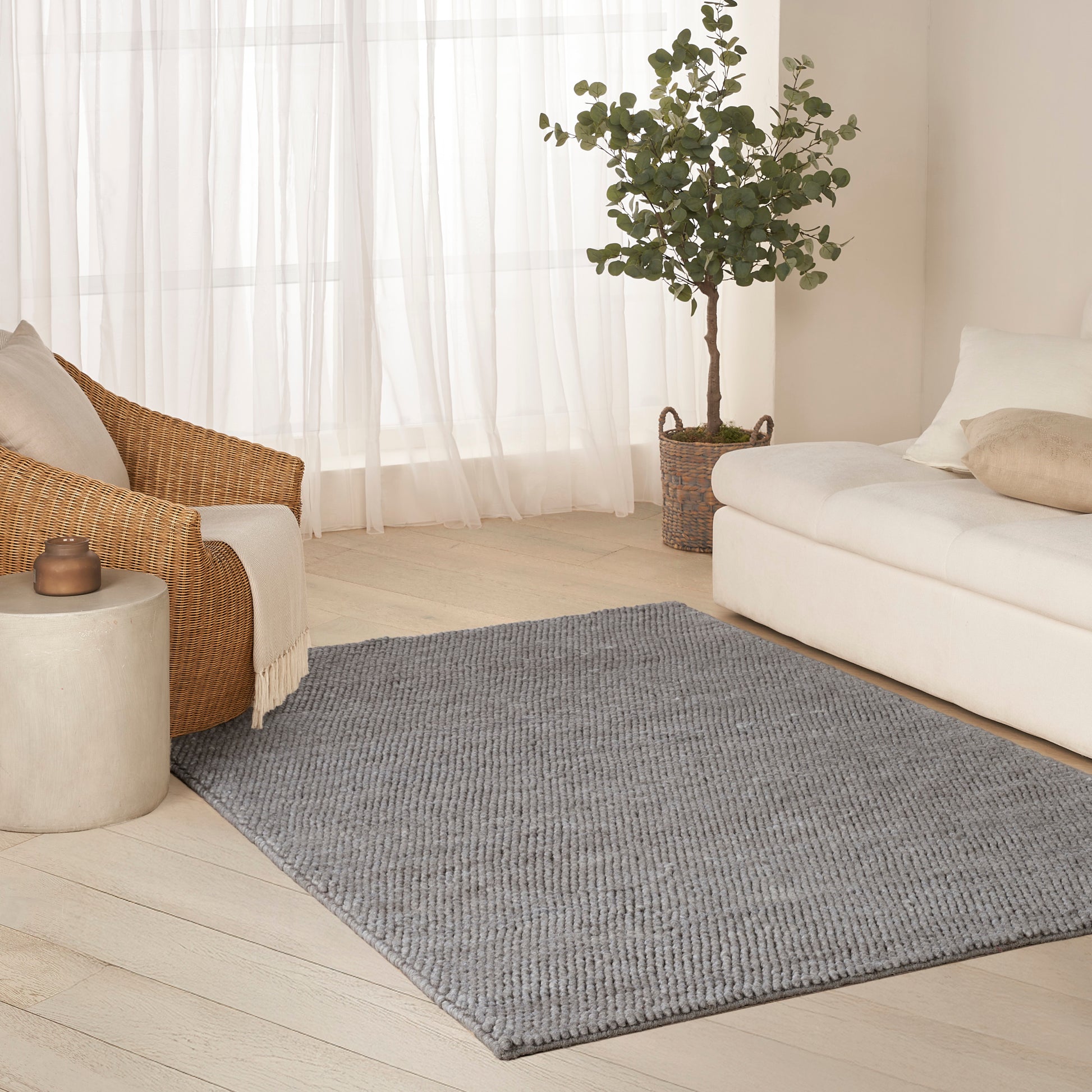 Light gray textured rug in a bright living room between a wicker chair and white sofa, with sheer curtains and a potted plant.