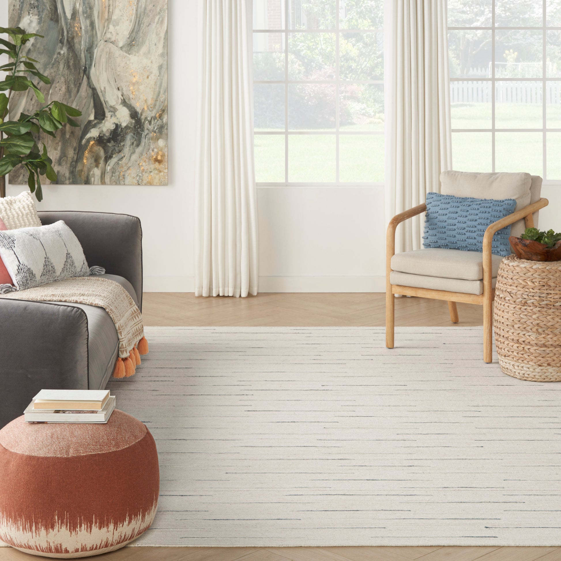 Cozy living room with beige rug, gray sofa, coral pouf, beige armchair with blue cushion, wicker side table, large window.