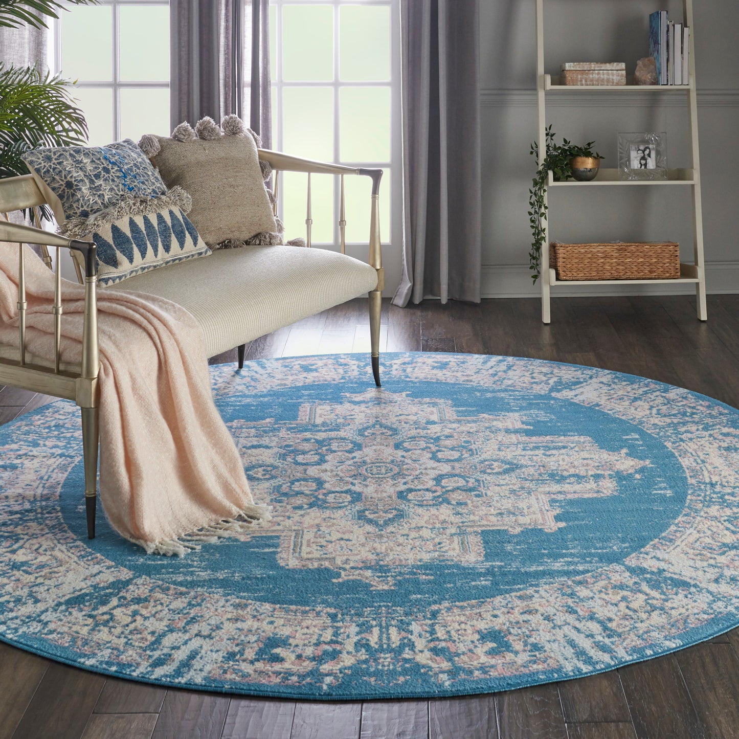 Round blue and ivory patterned rug on dark wood floor beside a cream metal-framed sofa with blue cushions and a pink throw