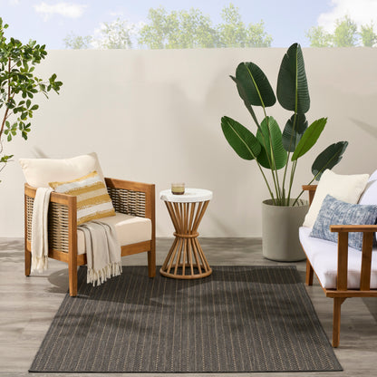 Outdoor patio rug with a dark striped pattern under wooden armchairs, beige cushions, a round side table, and a tall potted tropical plant.
