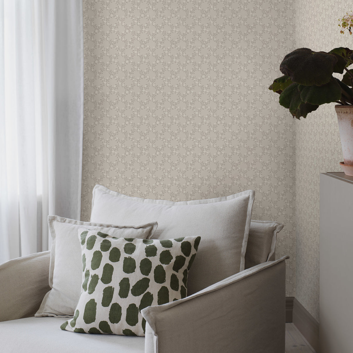 Living room corner with beige sofa & neutral cushions, against grayscale patterned wallpaper; green leaf-patterned pillow, sheer curtains & a potted plant on a cabinet.