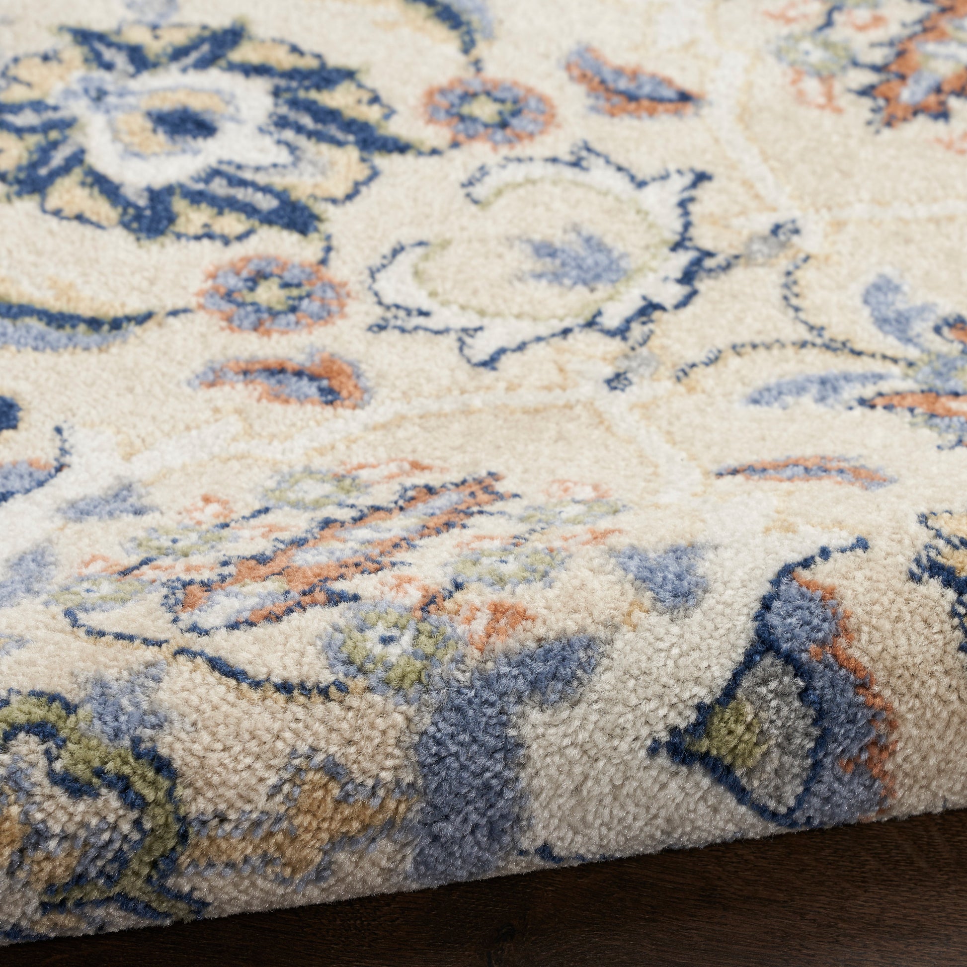 Cream floral patterned shag rug with blue, navy, orange and green accents on dark wood floor.