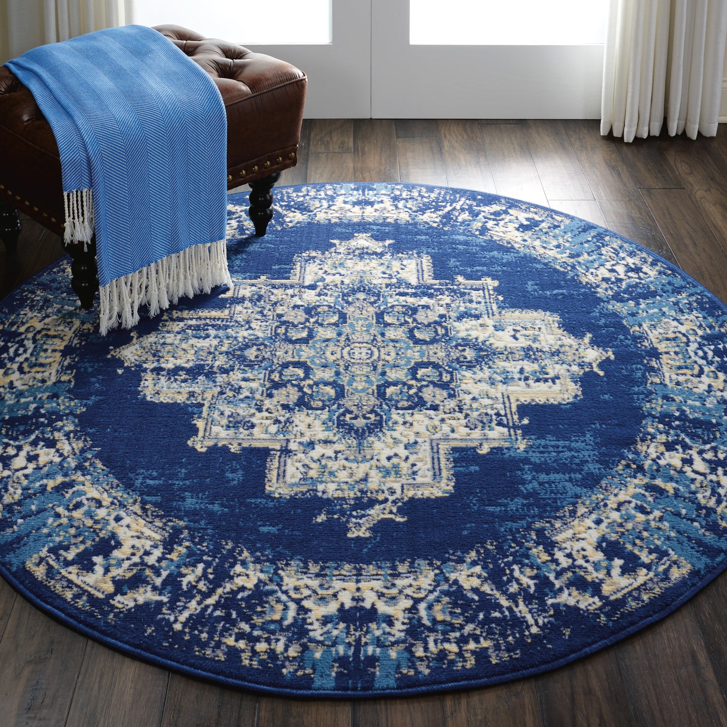 Round blue oriental rug with cream pattern on dark wood floor; tufted brown leather ottoman with blue fringed throw.