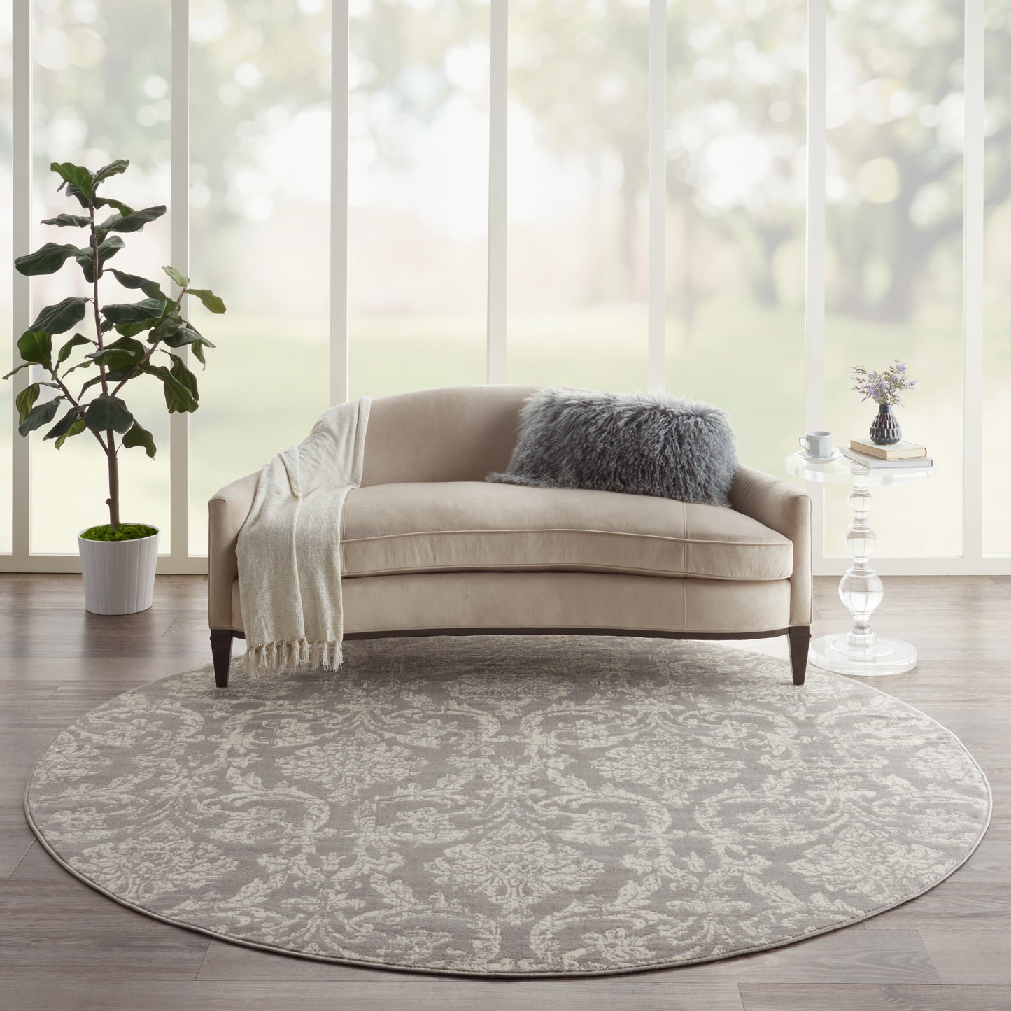 Beige curved sofa on round gray patterned rug with fluffy gray cushion, throw blanket in bright modern living room.