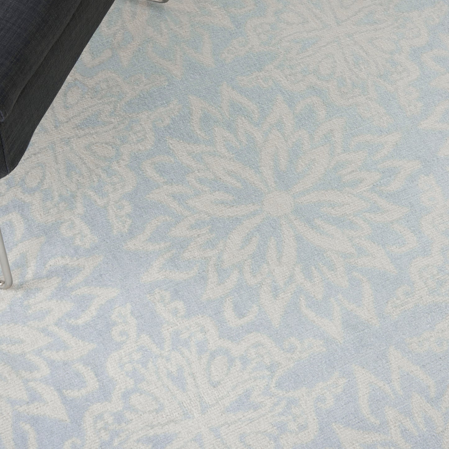 Light blue rug with large white snowflake/mandala pattern; soft textured surface with a partial black chair leg in the corner.