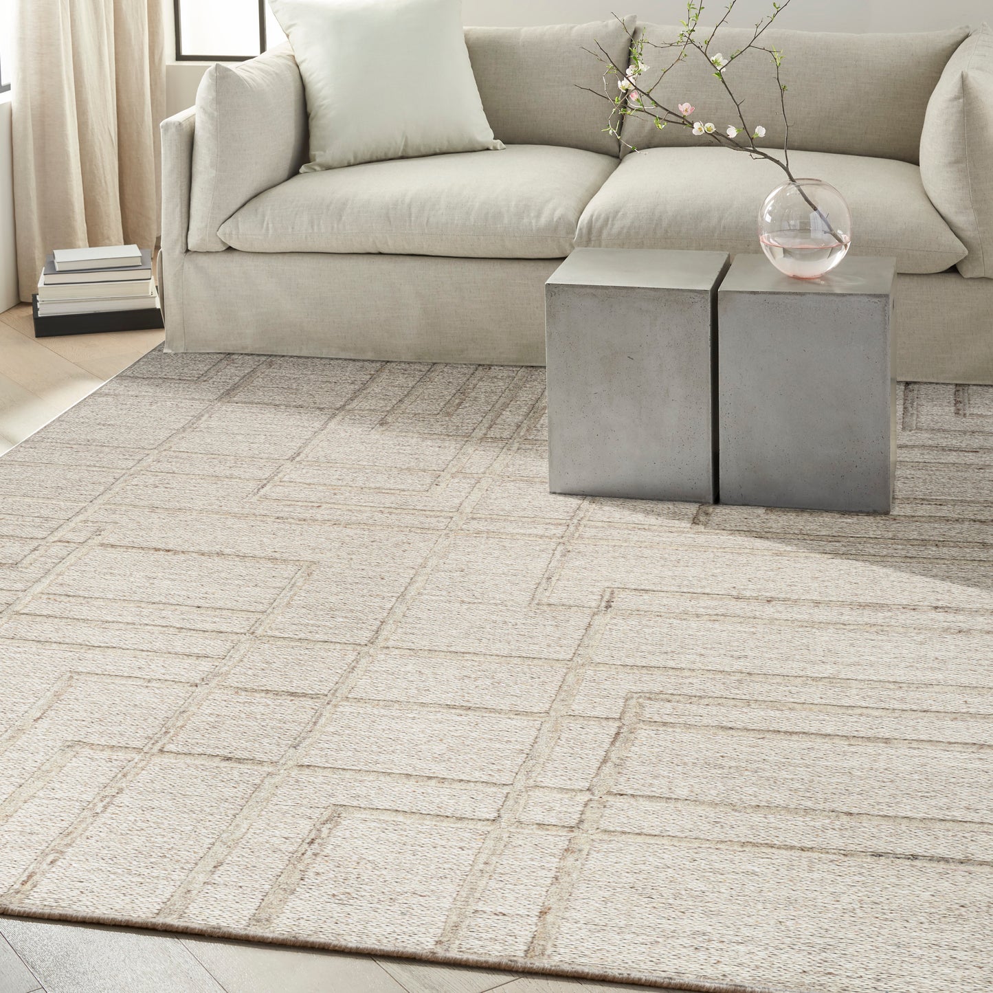 Beige fabric sofa and cream geometric area rug; two gray cube side tables with a glass vase and branch.