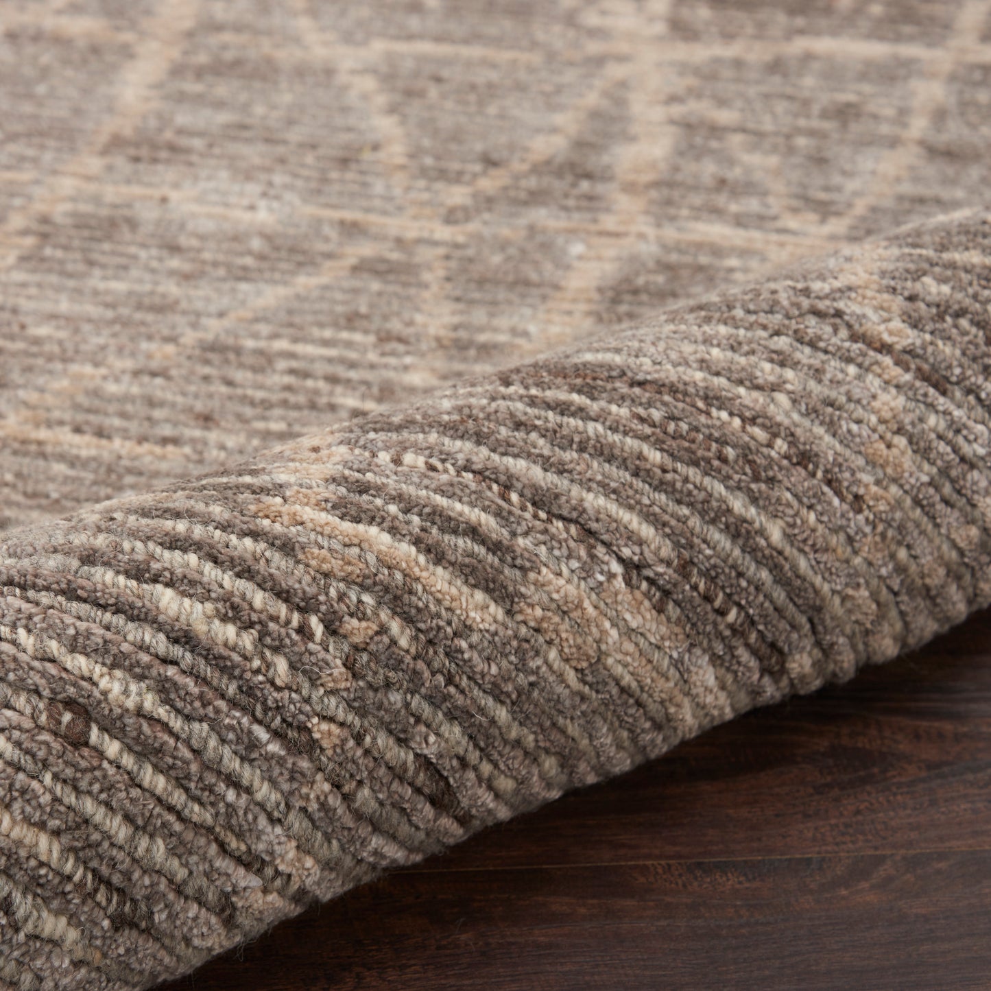 Beige-brown loop-pile area rug with geometric pattern, 8' x 10', rolled at edge on dark wood floor.
