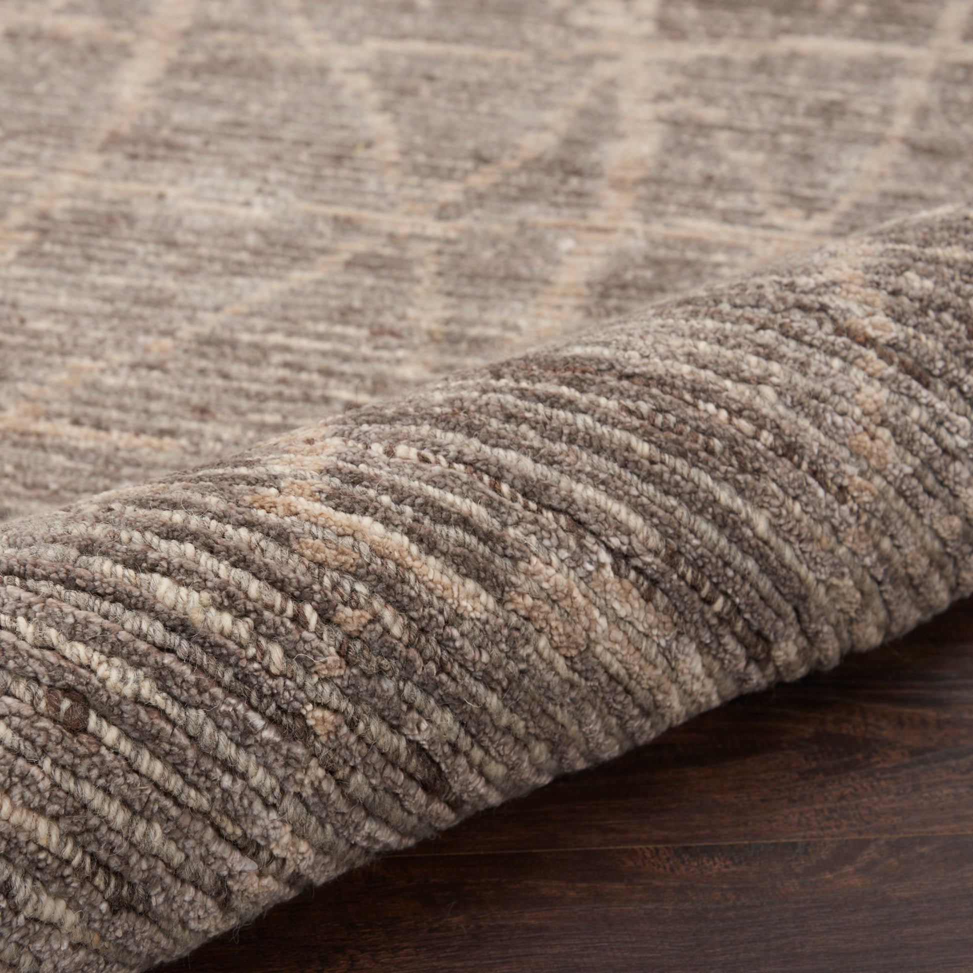 Beige-brown loop-pile area rug with geometric pattern, 8' x 10', rolled at edge on dark wood floor.