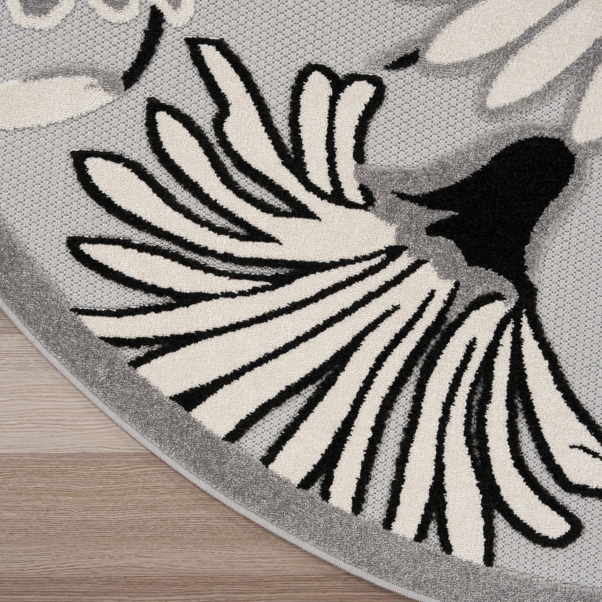 Round gray rug with bold black and cream floral petal motif, woven texture, gray edge, on wooden floor.