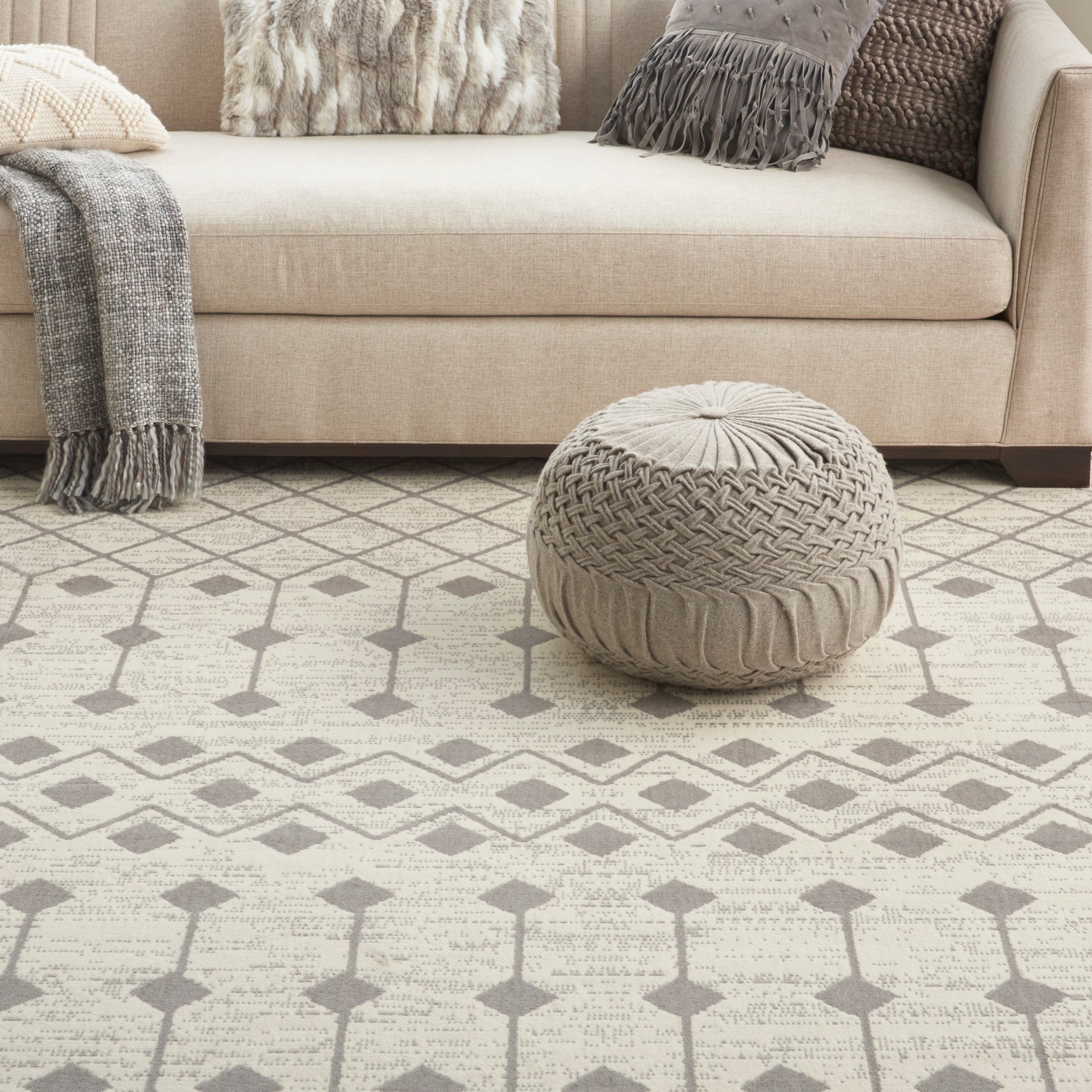 Beige sofa with textured pillows, a knit pouf ottoman, and a gray throw on a neutral geometric patterned rug.