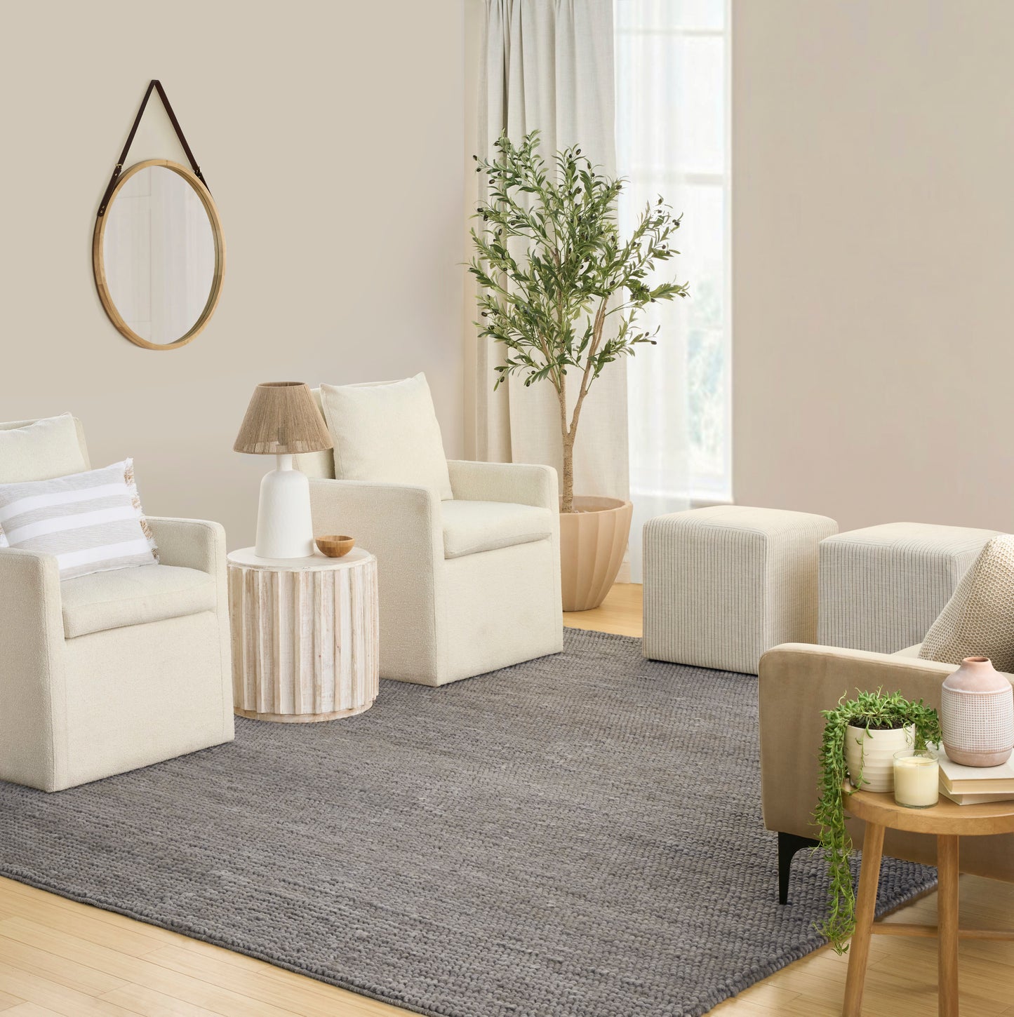 Neutral living room with cream armchairs, round mirror with leather strap, gray rug, potted tree, and wooden side table.