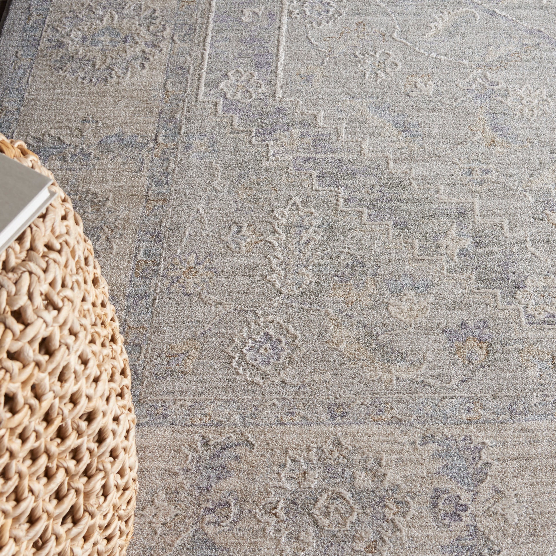 Beige and gray vintage-style rug with muted floral motifs; woven rattan pouf visible at left.