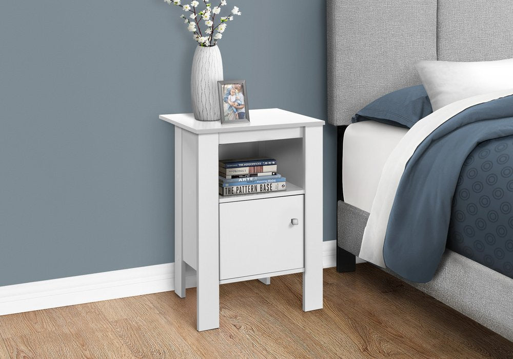 White wood end table with a shelf, vase of white flowers on top beside a gray upholstered bed with blue bedding.