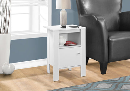White wood end table with shelf, lamp on top & red book, beside a gray leather armchair.