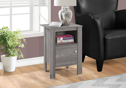 Gray weathered wood end table with lamp, open shelf with books, small drawer, beside black leather armchair.