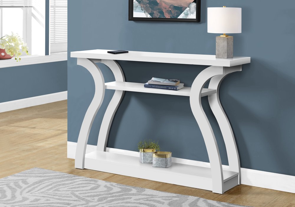 White wood console table with sculptural curved legs and two shelves on a blue wall, styled with lamp and books.