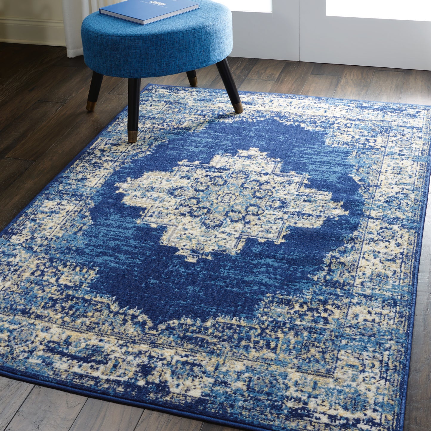 Blue distressed area rug with ivory central medallion on dark wood floor beside a blue round upholstered ottoman