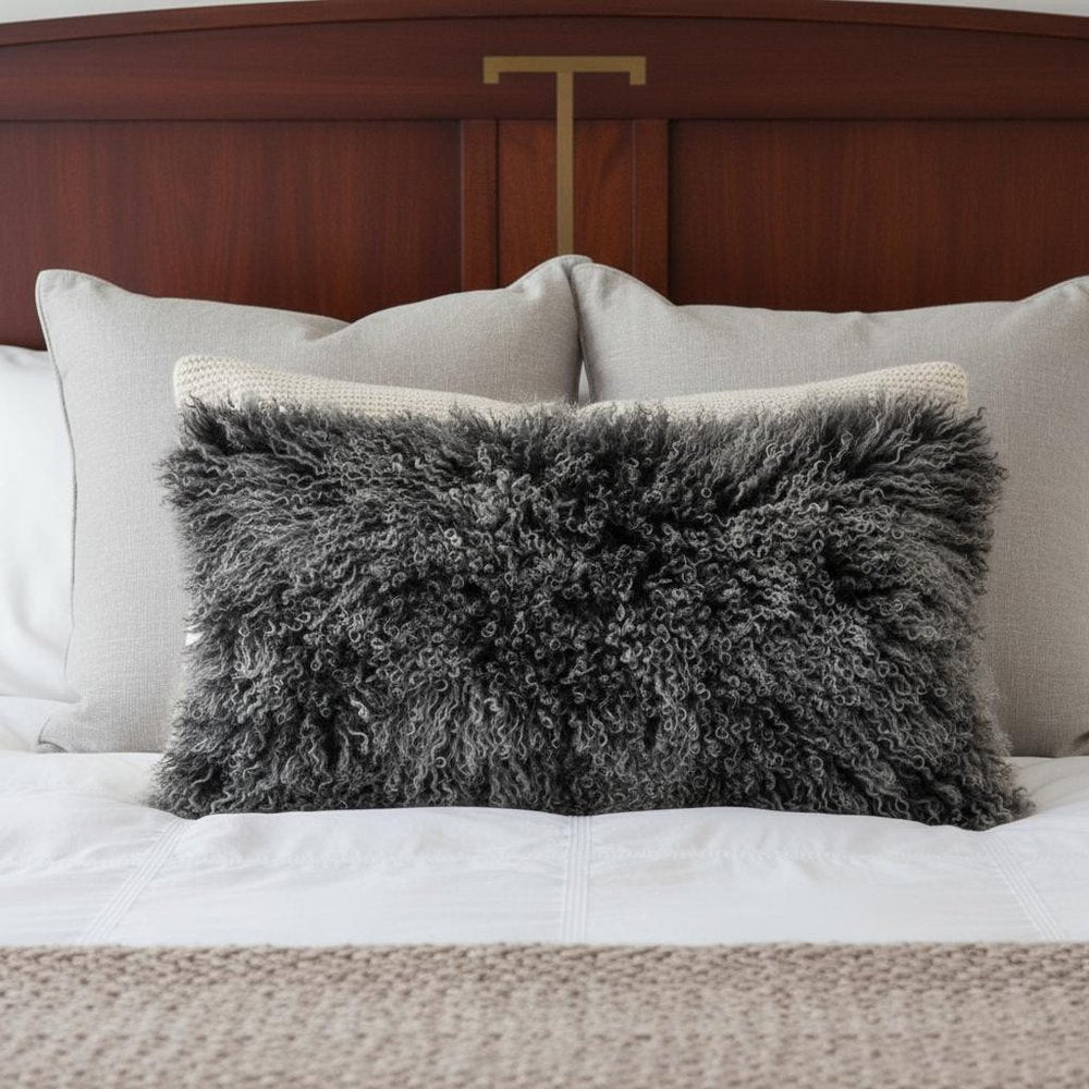 Rectangular 17" black genuine Tibetan lamb fur pillow with microsuede backing on a white bed with light-gray cushions and a wooden headboard.