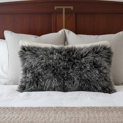 Rectangular 17" black genuine Tibetan lamb fur pillow with microsuede backing on a white bed with light-gray cushions and a wooden headboard.