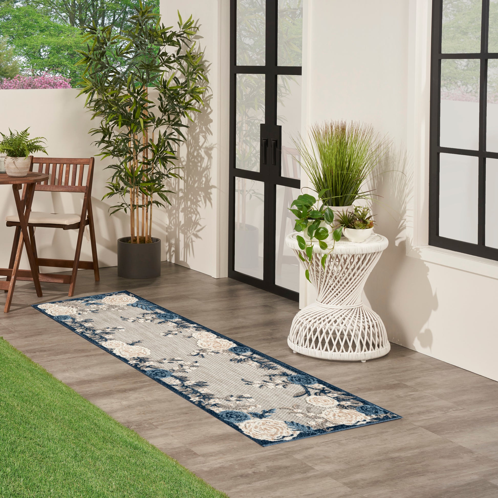 Blue-gray patterned runner rug on a wood floor beside a wicker plant stand and potted plants near glass doors.