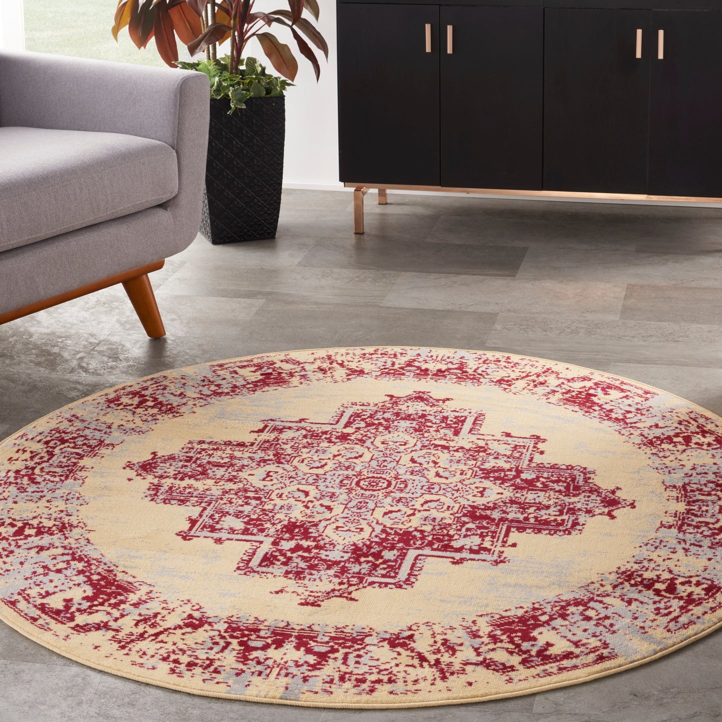Round beige rug with a red oriental medallion pattern in a modern living room; gray sofa, black cabinet, potted plant.