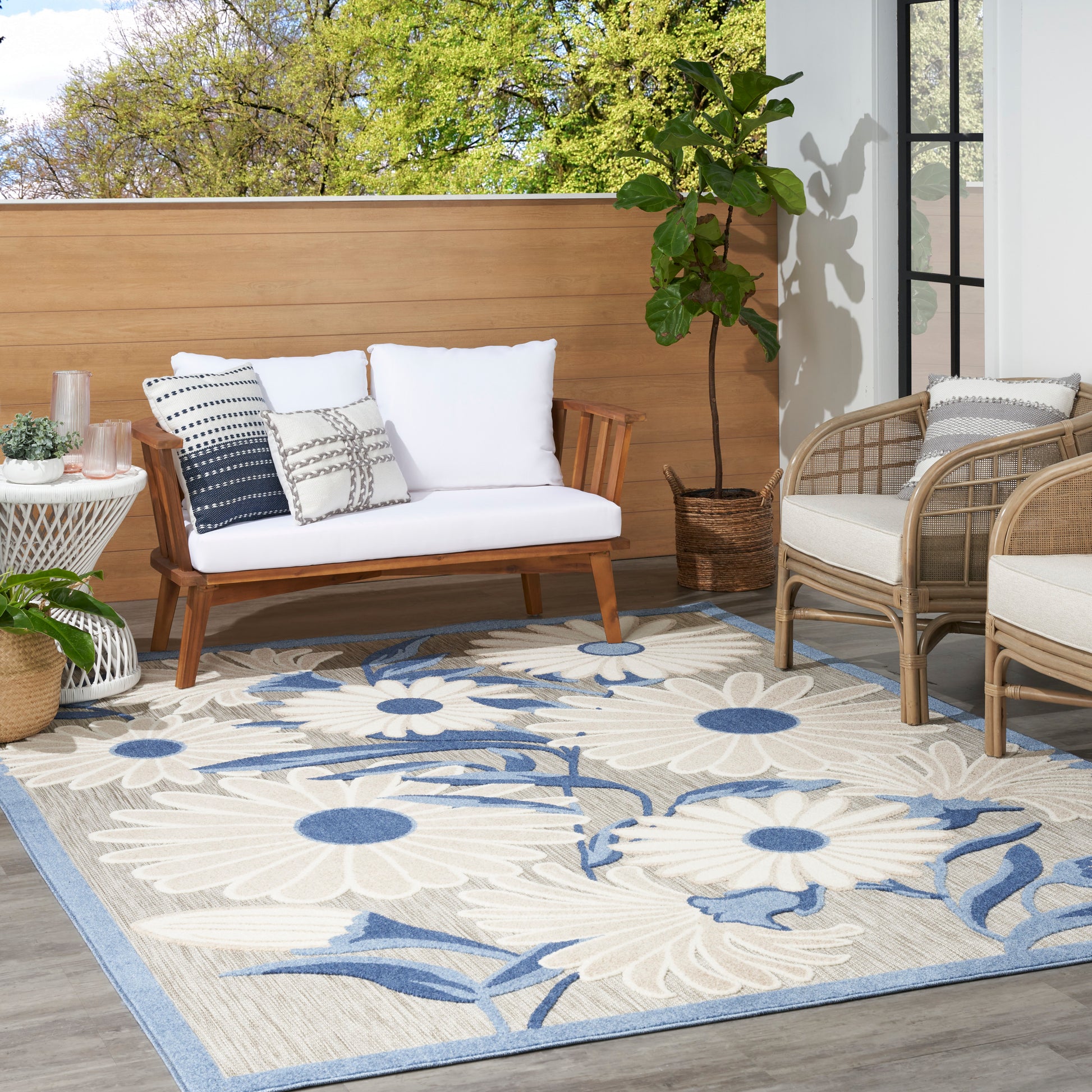 Outdoor patio with white cushioned wooden sofa, woven-rattan chairs, a large blue floral rug, and potted plants.