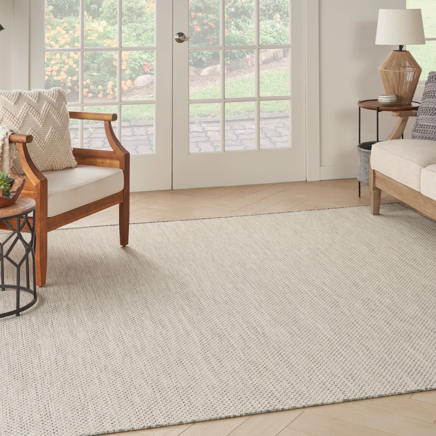 Beige woven area rug anchors a light wood living room with wooden armchairs, sofa, side table, and glass French doors.
