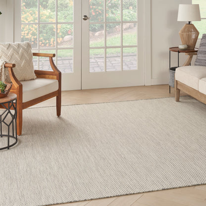 Beige woven area rug anchors a light wood living room with wooden armchairs, sofa, side table, and glass French doors.