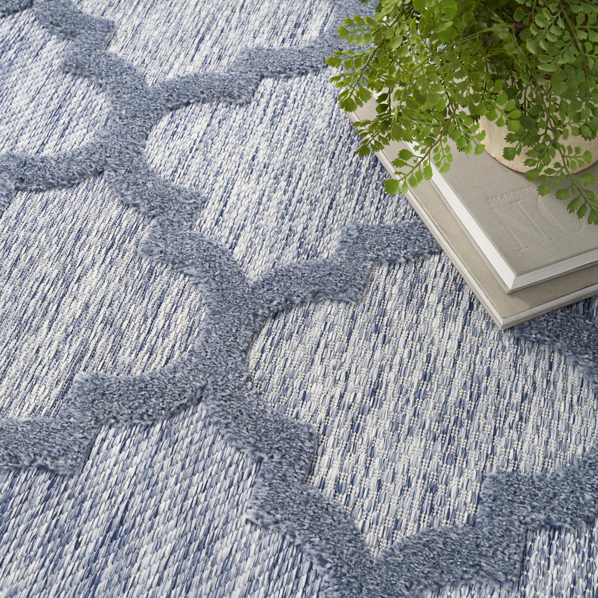 Blue-gray textured rug with a repeating trellis pattern; corner features a potted plant atop stacked books
