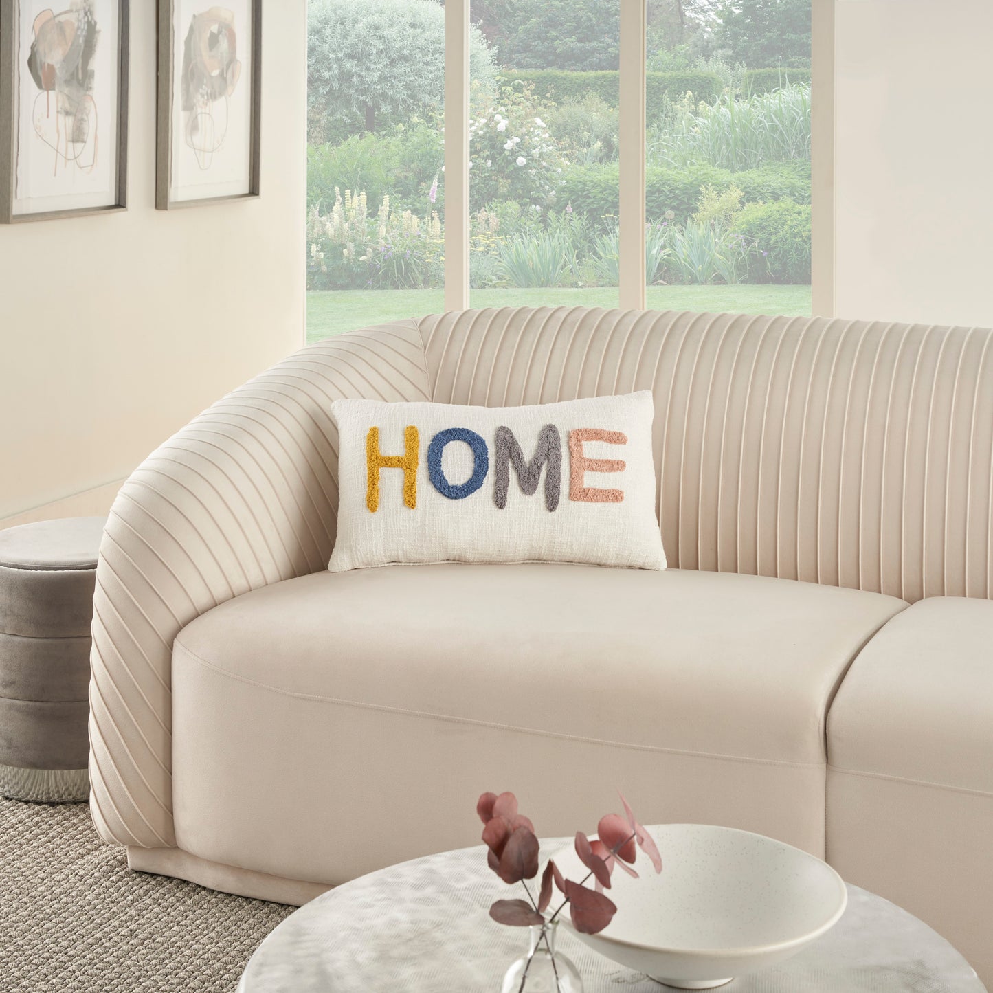Cream curved sofa with ribbed back, colorful 'HOME' embroidered pillow, round marble coffee table, soft neutral living room.