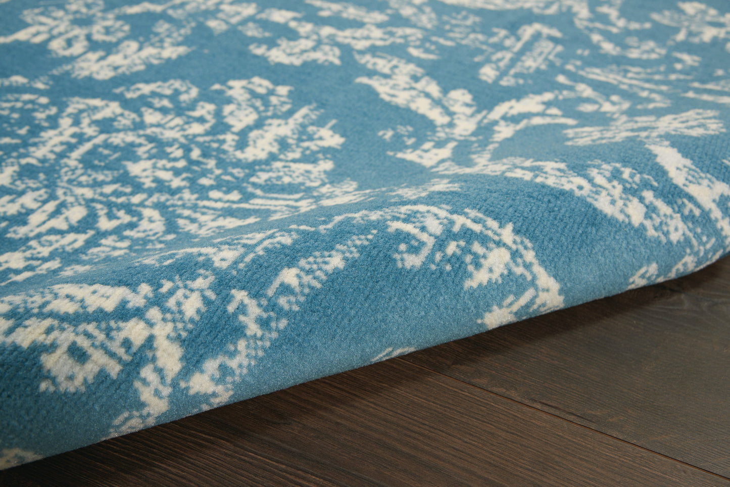 Blue area rug with white damask floral pattern, low-pile, rolled edge on dark wood floor.