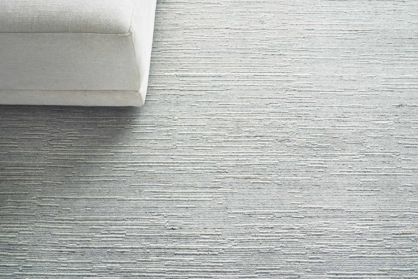 Light gray textured area rug with subtle horizontal stripes; beige sofa corner in a modern minimalist living room.