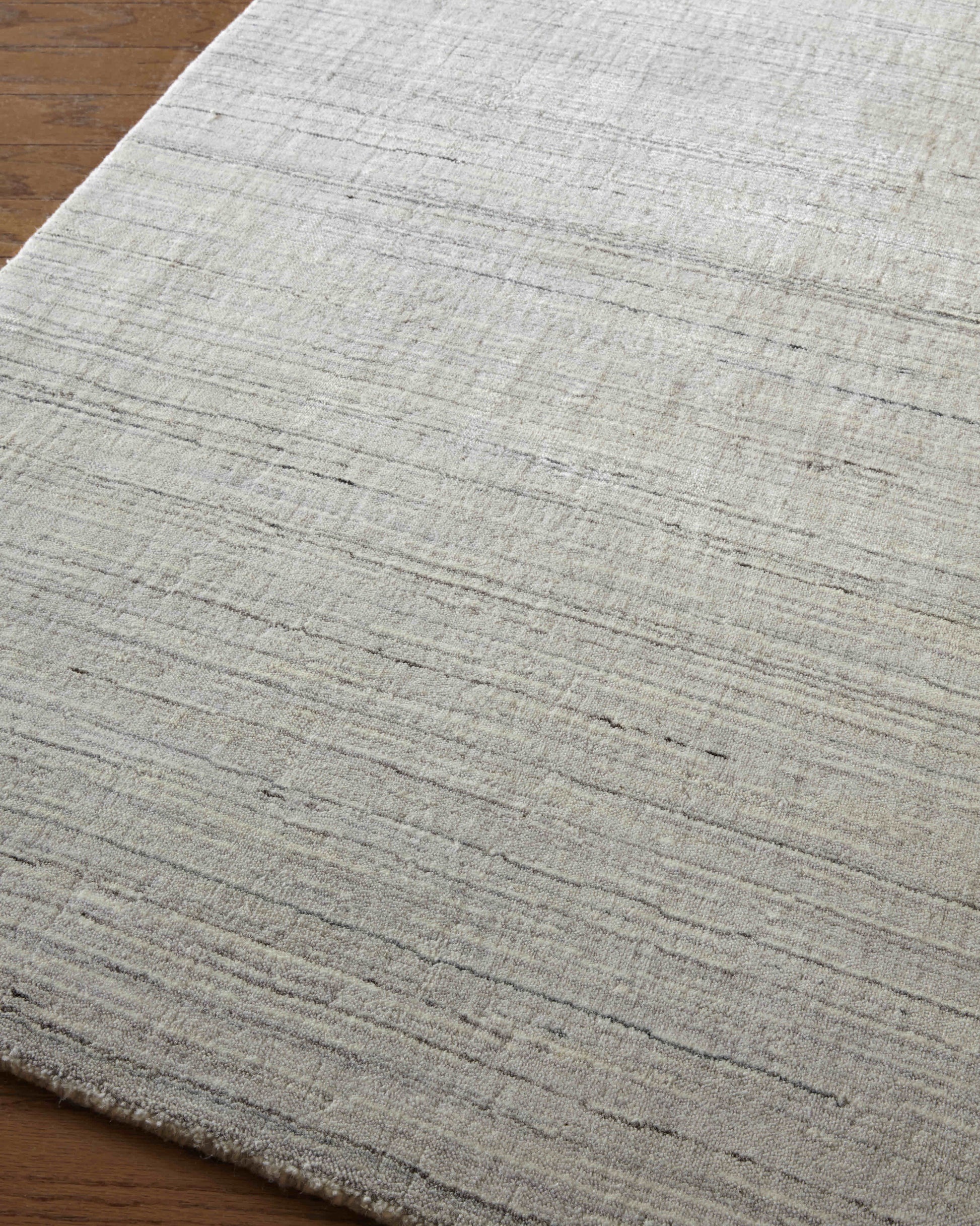 Light gray textured rug with subtle horizontal stripes and a fringed edge on a wood floor.