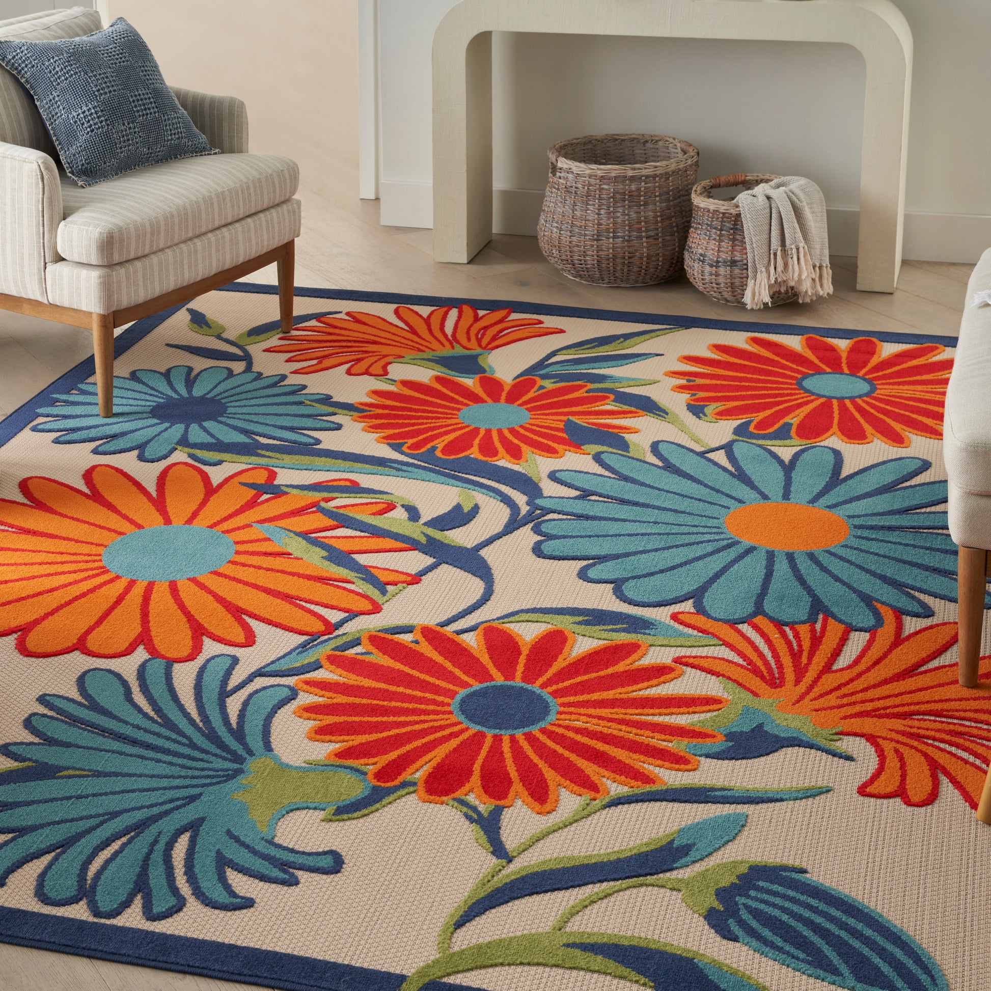 Beige floral area rug with large orange and teal petals and a blue border, 5' x 8', in a cozy living room with a sofa and wicker baskets.