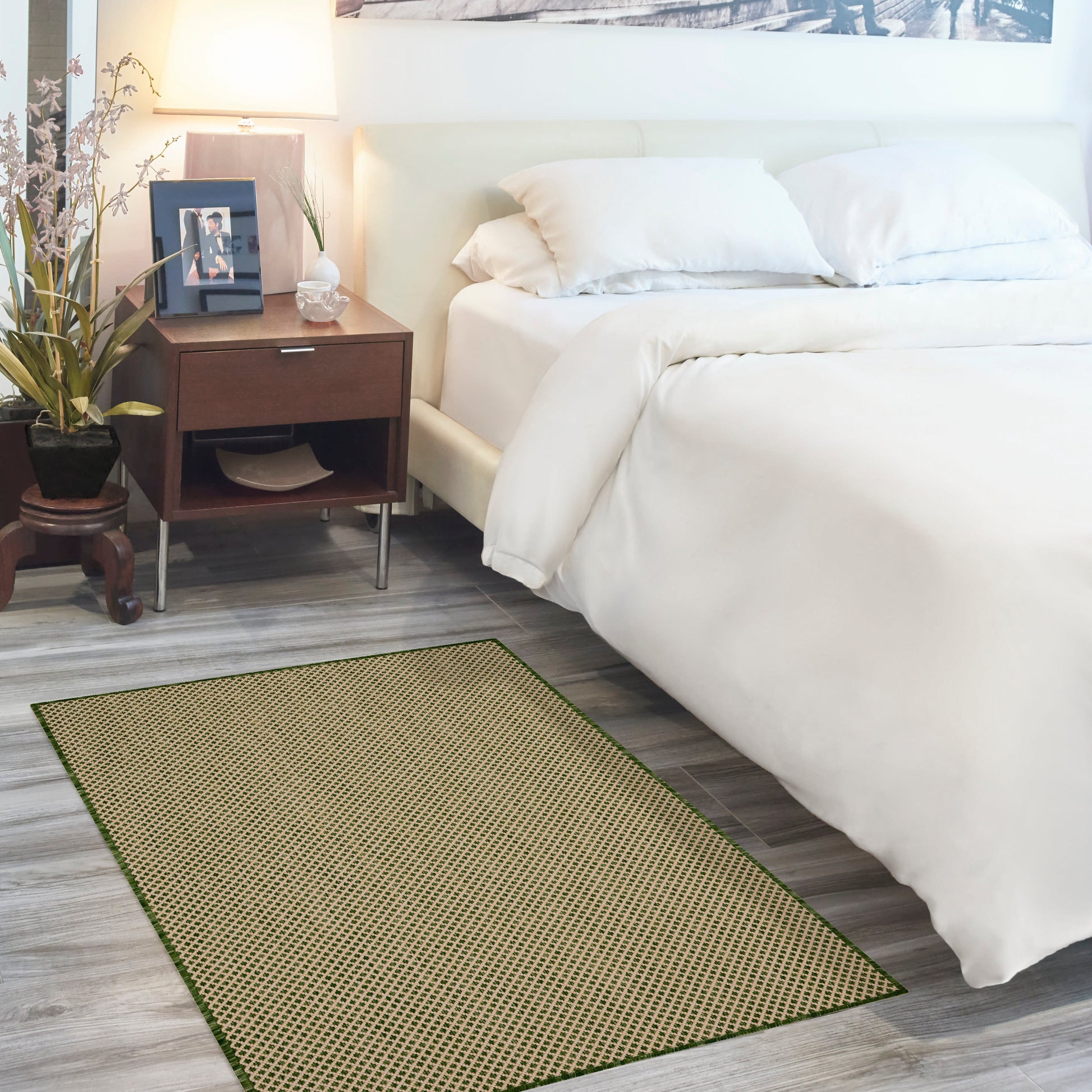 White bed with crisp duvet, dark wood nightstand, potted plants, and a green woven rug on gray wood floor.