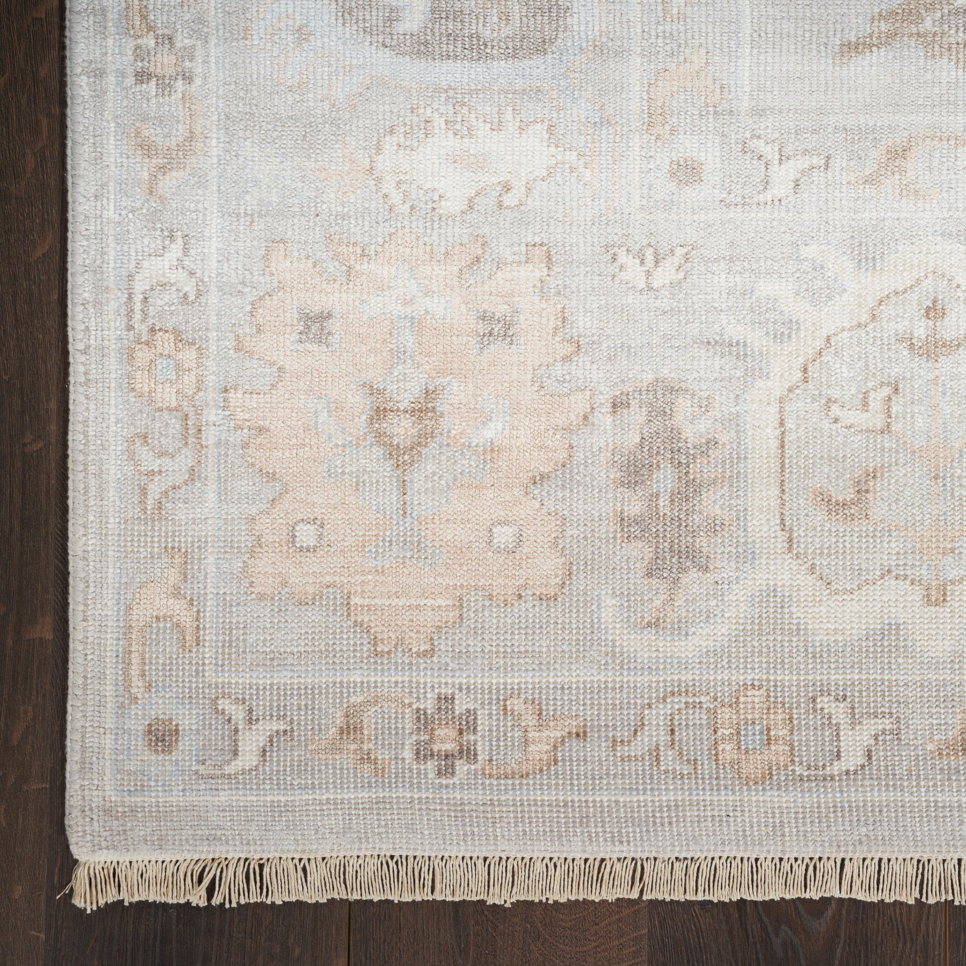 Muted beige, gray and cream oriental rug with floral medallion border and fringed edge on dark wood floor.